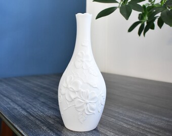 May include: A white ceramic vase with a floral relief pattern. The vase has a slender neck and a flared base. The background includes a blue wall, a white wall, and green leafy branches. The vase is approximately 10 inches tall.