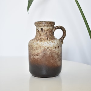 May include: A brown and white ceramic vase with a speckled glaze and a handle. The vase has a rounded body and a narrow neck.