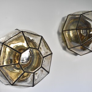 May include: Two geometric wall sconces with a clear, faceted glass design and a gold-toned interior. The sconces are mounted on a white wall, showcasing their unique, decorative shape and light-reflecting qualities. The metal frames are black.