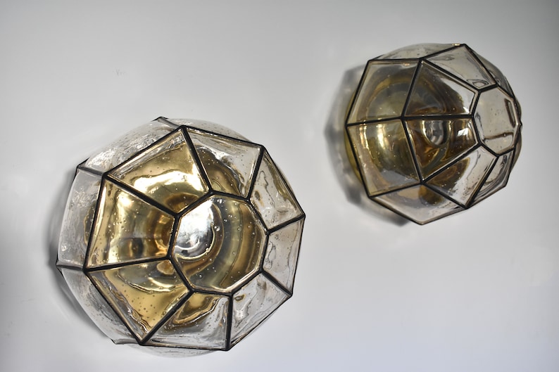 May include: Two geometric wall sconces with a clear, faceted glass design and a gold-toned interior. The sconces are mounted on a white wall, showcasing their unique, decorative shape and light-reflecting qualities. The metal frames are black.
