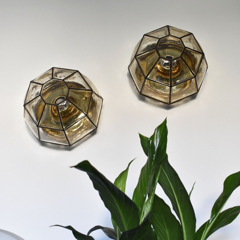 May include: Two geometric, amber-colored glass wall sconces with black metal frames are mounted on a white wall. The sconces have a faceted design. Green plant leaves are visible in the foreground.