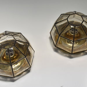May include: Two geometric, dome-shaped wall sconces. Each sconce features a clear, faceted glass shade with a black metal frame. The interior has a warm, amber glow. The sconces are mounted on a white wall.