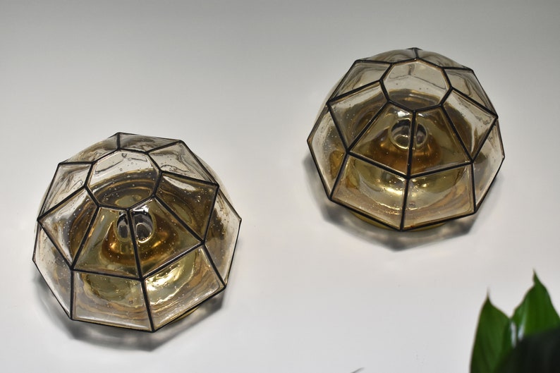 May include: Two geometric, dome-shaped wall sconces. Each sconce features a clear, faceted glass shade with a black metal frame. The interior has a warm, amber glow. The sconces are mounted on a white wall.