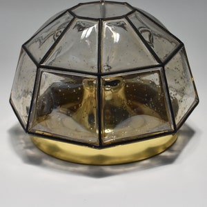 May include: A decorative ceiling light fixture with a geometric, domed shade. The shade is made of clear, faceted glass with black trim, and sits atop a gold-colored base. The glass has a slightly amber tint.