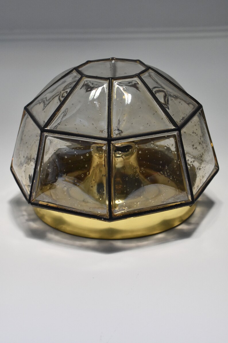 May include: A decorative ceiling light fixture with a geometric, domed shade. The shade is made of clear, faceted glass with black trim, and sits atop a gold-colored base. The glass has a slightly amber tint.