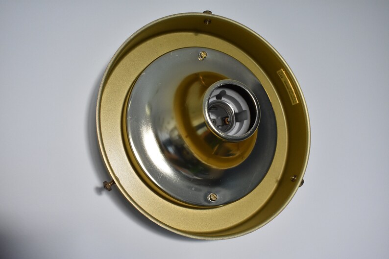 May include: A gold-colored ceiling light fixture with a central silver-colored bulb socket. The fixture has a circular design with a brass-colored outer ring and is mounted on a white surface.