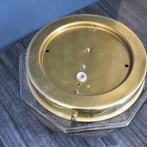 May include: A vintage brass clock face with a clear, octagonal glass base. The brass face has several small holes and a central keyhole. The clock sits on a dark gray, textured surface.