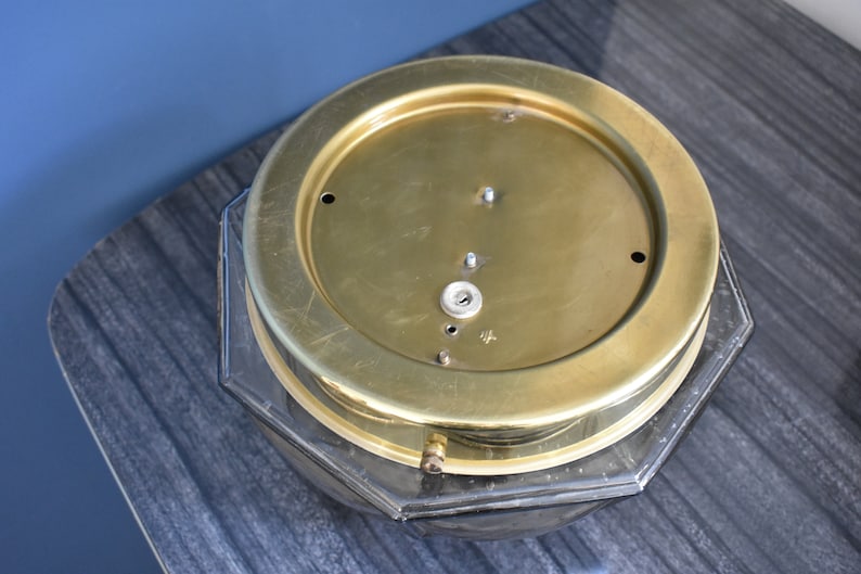 May include: A vintage brass clock face with a clear, octagonal glass base. The brass face has several small holes and a central keyhole. The clock sits on a dark gray, textured surface.