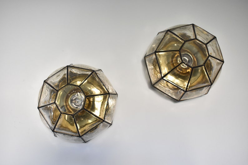 May include: Two geometric-shaped wall sconces. Each sconce features a multifaceted, clear glass design with a gold-toned interior. The glass panels are outlined with black metal. The sconces are mounted on a white wall, providing a modern lighting fixture.