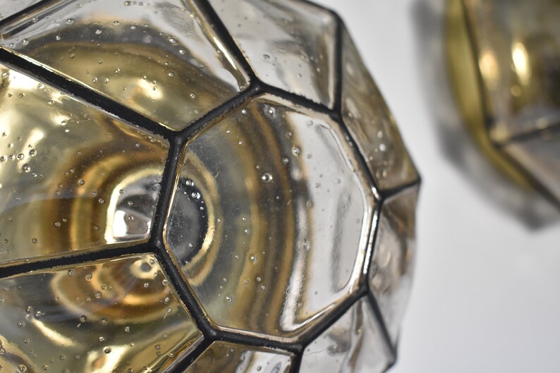 May include: Close-up of two geometric glass objects with a metallic finish. The objects have a multifaceted design with black outlines. The glass has a reflective surface with small bubbles, and the interior has a golden hue.