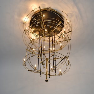 Könnte beinhalten: Ein goldfarbener Kronleuchter mit mehreren runden Ebenen und einem schwarzen Zentrum. Der Kronleuchter hat viele kleine Lichter, die eingeschaltet sind.