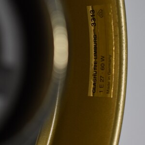May include: Close-up of a gold-colored object with a curved shape. A rectangular sticker is affixed to the object, displaying the text "GLASHUTTE-LIMBURG 3313 1 E 27 60 W Made in Germany". The object has a glossy finish.