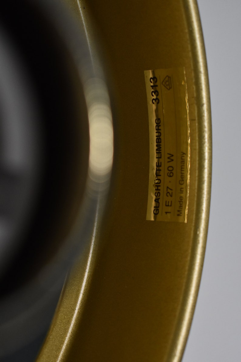 May include: Close-up of a gold-colored object with a curved shape. A rectangular sticker is affixed to the object, displaying the text "GLASHUTTE-LIMBURG 3313 1 E 27 60 W Made in Germany". The object has a glossy finish.