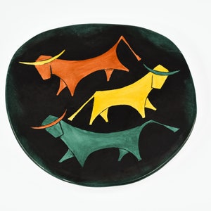 May include: A black ceramic plate with a unique design featuring three stylized bulls. The bulls are in shades of orange, yellow, and green, each with yellow horns. The plate has an irregular, organic shape.