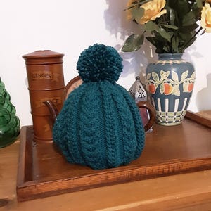 Könnte beinhalten: Ein tealfarbener, gestrickter Teekannenwärmer mit einem weißen Bommel oben. Der Wärmer liegt auf einer Holzoberfläche, aus der ein Teekannen-Ausguss herausragt.