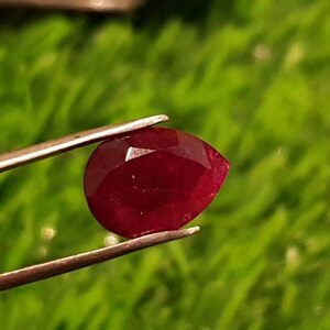 May include: A deep red pear-shaped gemstone held by tweezers against a green background.