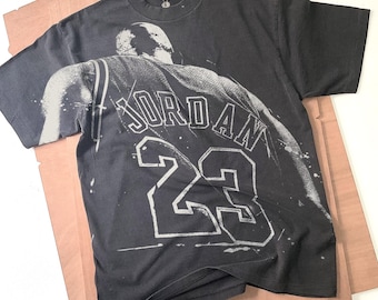 Op de afbeelding: Donkergrijs t-shirt met een verweerde afbeelding van een basketbalshirt met de naam "JORDAN" en het nummer "23" in wit. Het shirt heeft een vintage, versleten uitstraling.