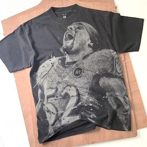 Puede incluir: Camiseta gris oscuro con un gráfico desgastado de un jugador de fútbol americano con el uniforme de los Ravens, con el número 52. La palabra "Art" está en un círculo en la camiseta. La camiseta está sobre una superficie de papel marrón.