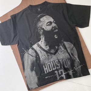 Könnte beinhalten: Schwarzes T-Shirt mit einem Graustufenbild eines Basketballspielers. Der Spieler trägt ein Trikot mit "HOUSTON" und der Nummer 13. Das Nike-Logo ist ebenfalls sichtbar. Das T-Shirt hat einen Rundhalsausschnitt.