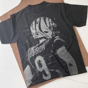 May include: Black t-shirt with a graphic print of a football player wearing a helmet and jersey with the number 9 and the name "Burrow" on the back.