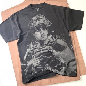 Könnte beinhalten: Dunkelgraues T-Shirt mit einem Graustufenbild eines Baseball-Catchers. Die Grafik zeigt den Helm, den Brustschutz und einen konzentrierten Gesichtsausdruck. Das kurzärmelige Shirt hat einen Rundhalsausschnitt.