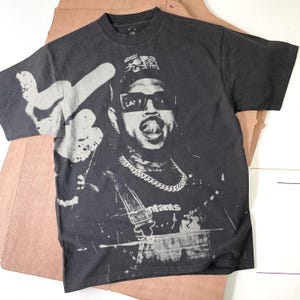 May include: Black graphic t-shirt featuring a stylized image of a person wearing sunglasses, a hat, and a chain. The design includes the word "Infants" and a hand gesture. The shirt has a vintage, distressed look.