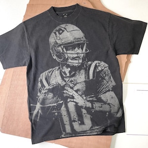 Puede incluir: Camiseta negra con una imagen en escala de grises de un jugador de fútbol americano con casco y camiseta con el número 10. El jugador sostiene un balón. La camiseta tiene un aspecto vintage y desgastado.