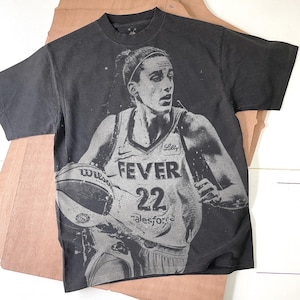 May include: Dark grey t-shirt with a grayscale image of a basketball player holding a basketball. The player wears a jersey with "FEVER" and the number "22". The shirt also has the word "salesforce" printed on it.