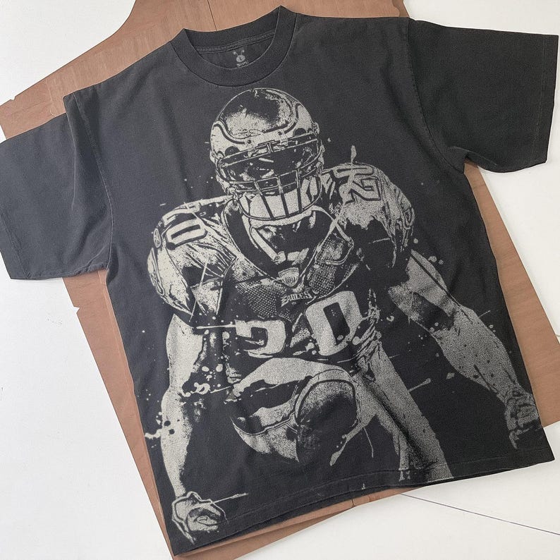 May include: Black t-shirt featuring a grayscale image of a football player in action. The player is wearing a helmet and uniform with the number 20 visible. The word "Eagles" is also visible on the uniform. The shirt is laid on a brown surface.