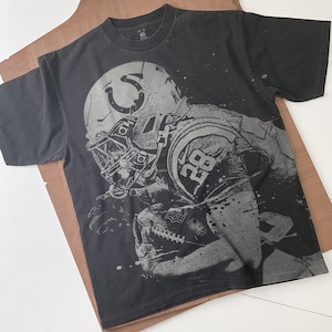Könnte beinhalten: Schwarzes T-Shirt mit einer grauen Grafik eines American-Football-Spielers mit Helm, der einen Football hält. Das Trikot des Spielers zeigt die Nummer 28. Das Logo der Indianapolis Colts ist auf dem Helm zu sehen.
