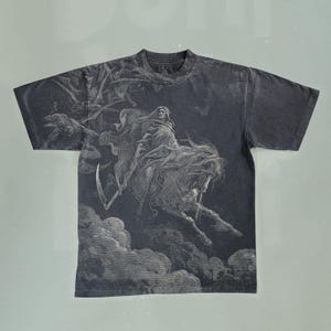Peut inclure: T-shirt gris foncé avec une illustration détaillée d'une figure vêtue d'une robe à cheval, tenant une faux, sur fond nuageux. Le graphisme est en nuances de gris et de blanc, créant une esthétique vintage. Le t-shirt a un col rond classique et des manches courtes.