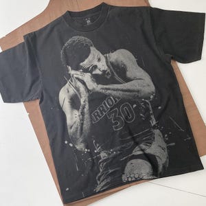 Peut inclure: T-shirt noir avec une image en niveaux de gris d'un joueur de basket-ball portant un maillot des Warriors avec le numéro 30. Le joueur est représenté avec les mains près du visage. Le t-shirt est sur une surface marron.