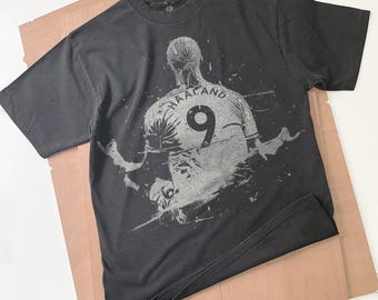 May include: Dark gray t-shirt featuring a graphic of a soccer player's back with the name "HAALAND" and the number "9" in white. The shirt is on a light brown surface.