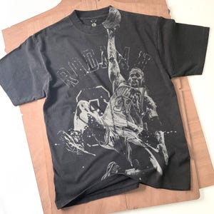 May include: Dark grey t-shirt with a black and white graphic of a basketball player, with the name "RODMAN" and the number "91". The shirt is displayed on a brown surface.