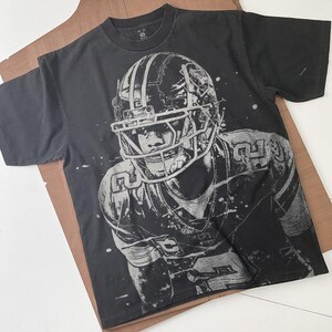 May include: Black t-shirt featuring a grayscale illustration of a football player in a helmet and uniform. The graphic has a distressed, splattered effect. The shirt is laid flat on a brown surface.