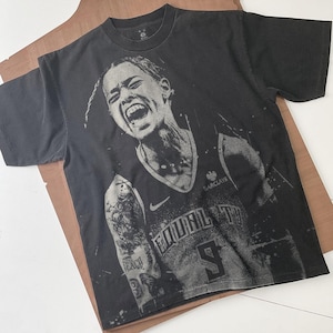 May include: Black t-shirt featuring a grayscale image of a basketball player with an open mouth. The shirt has a Nike logo and the word "Brooklyn" on the jersey. The player's jersey number is 9.