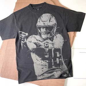 May include: Black t-shirt featuring a gray graphic of a football player in a helmet and uniform. The player's number 10 is visible on the jersey. The shirt has a crew neck and short sleeves. The background is a light brown surface.