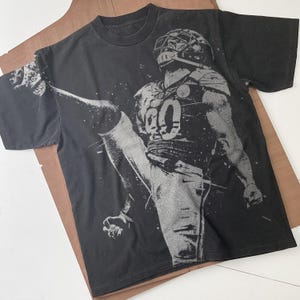 May include: Black t-shirt with a silver graphic print of a football player kicking a ball. The player is wearing a jersey with the number "80" on it.