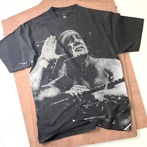 Puede incluir: Camiseta negra con una imagen en escala de grises de un luchador. La imagen muestra la cara y la parte superior del cuerpo del luchador, con la mano levantada. La camiseta tiene cuello redondo y mangas cortas. El luchador lleva una cinta en la cabeza.