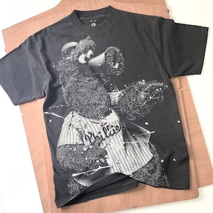 Puede incluir: Camiseta gris oscuro con un gráfico de un oso con una camiseta de béisbol de los Phillies. La camiseta es blanca con rayas azules y la palabra "Phillies". El oso sostiene un bate de béisbol.