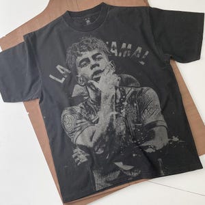 May include: Black t-shirt featuring a grayscale image of a soccer player with the text "LA LAMAL". The shirt has a crew neck and short sleeves. The graphic is distressed, giving the shirt a vintage look.