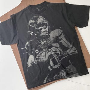 May include: Black t-shirt featuring a grayscale image of a football player in a helmet, holding a football. The player's jersey number is 10. The shirt is laid flat on a brown surface.