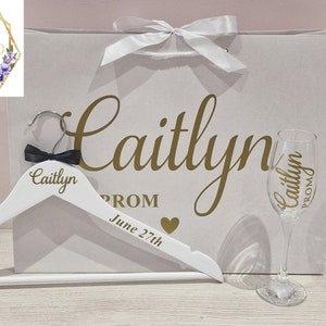 May include: White personalised hanger with the name Caitlyn and the word Prom, a white gift bag with the name Caitlyn and the words Prom and June 27th, and a clear glass champagne flute with the name Caitlyn and the word Prom.