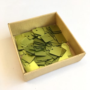 Puede incluir: Una caja de cartón llena de una gran cantidad de lentejuelas cuadradas de color verde metálico. Las lentejuelas tienen un pequeño agujero en el centro.
