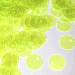 May include: A large pile of bright yellow, round sequins with a hole in the center. The sequins are scattered on a white surface.