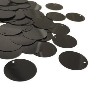 May include: A pile of black round sequins with a hole in the center. The sequins are scattered on a white background.
