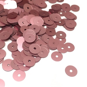 Puede incluir: Un montón de lentejuelas de color oro rosa, esparcidas sobre un fondo blanco. Las lentejuelas son redondas y tienen un agujero en el centro.