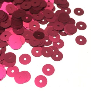 May include: A pile of loose, round, flat, metallic sequins in a deep pink color. The sequins are scattered on a white surface.