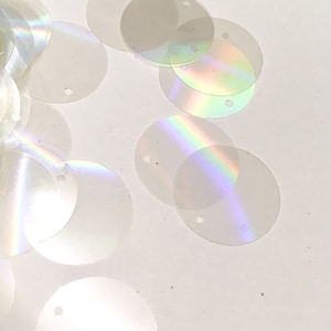 Peut inclure: Un tas de paillettes rondes blanches irisées avec un trou au centre. Les paillettes sont éparpillées sur une surface blanche.