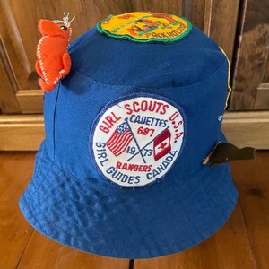 May include: A blue bucket hat with various patches and an orange felt creature. Patches include "Pack Holiday" and a Girl Guides emblem with US and Canadian flags, dated 1973. The hat is made of a durable fabric.